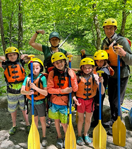 Children with two adults wearing river safety gear and holding paddles.